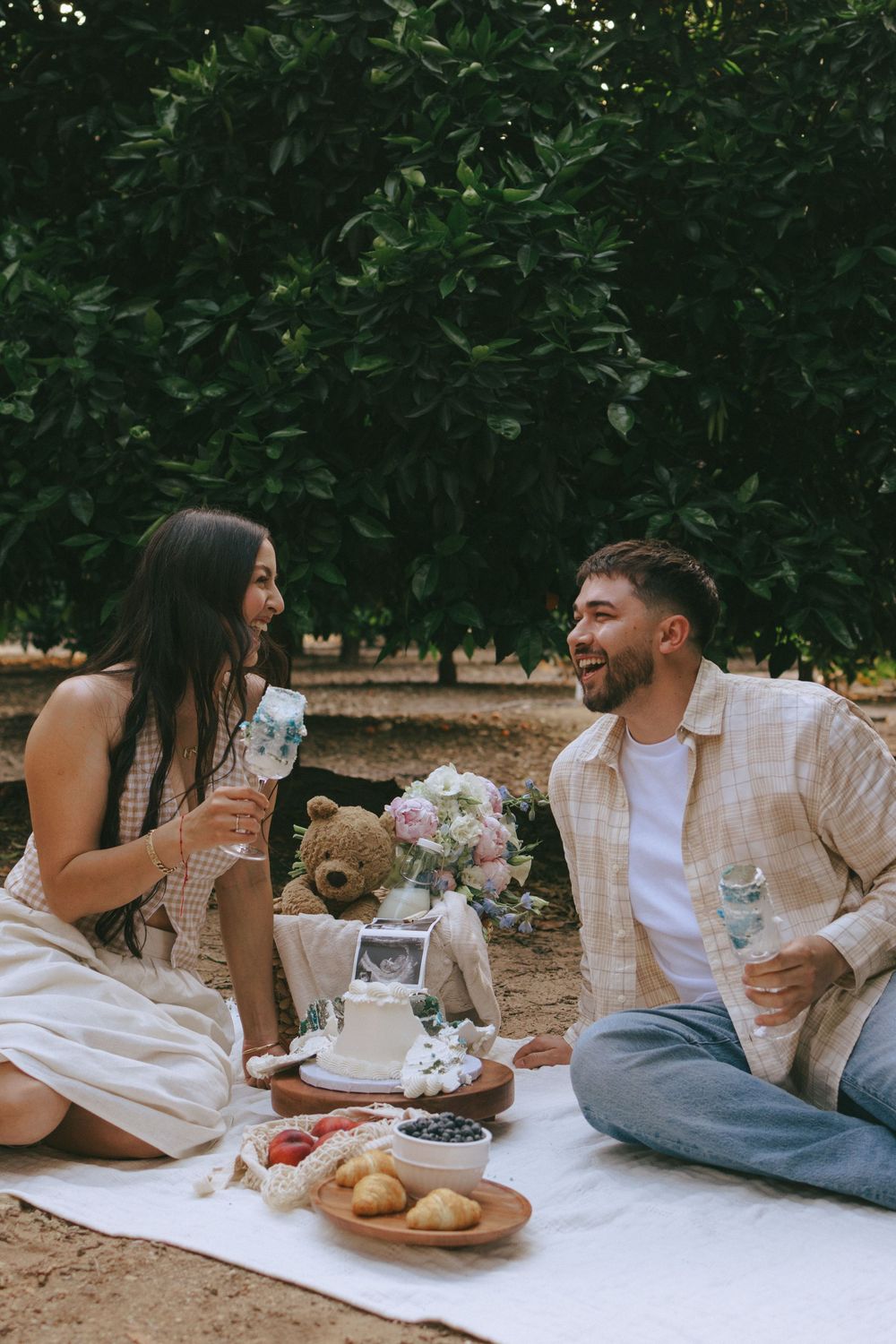 Collage showing couple sitting on picnic blanket outdoors sharing meal and drinks under green trees.