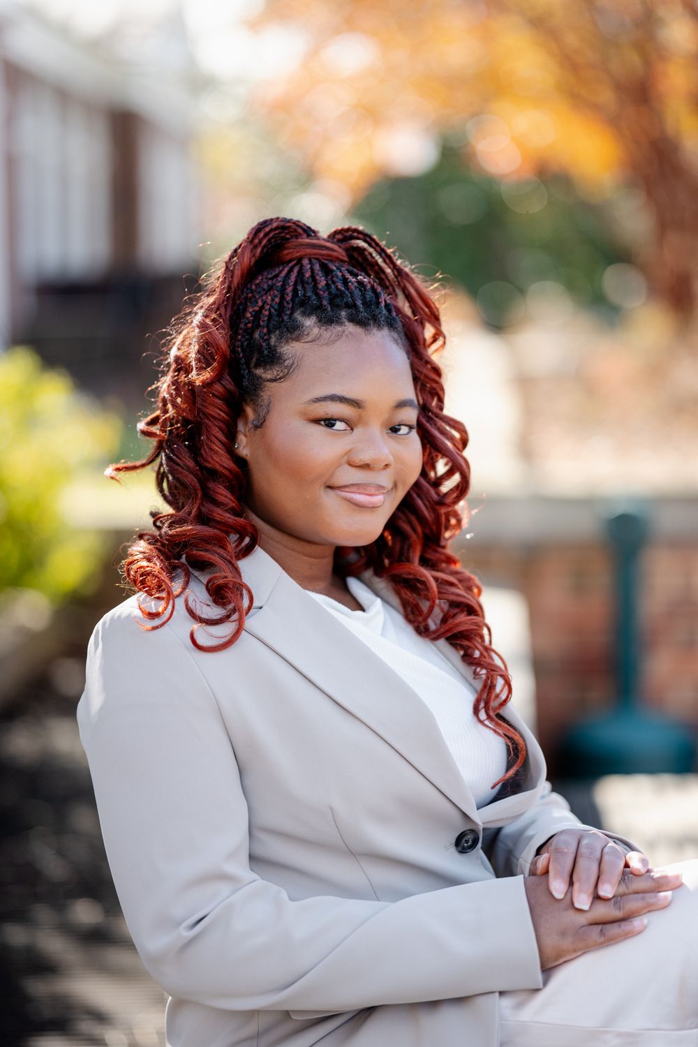 Young Black woman with red braided hair wearing a beige blazer, smiling outdoors in autumn setting.
