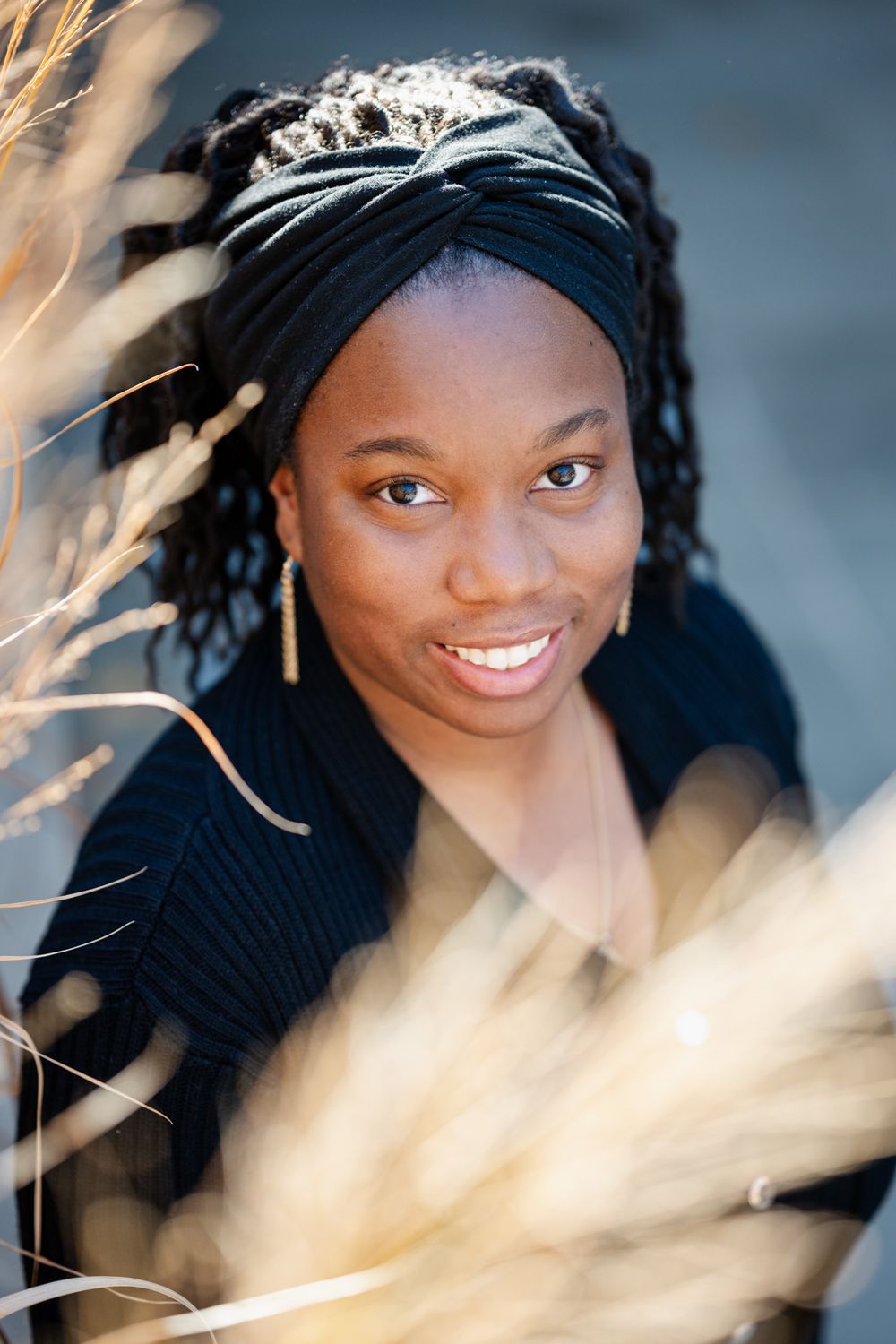 Young Black woman smiling outdoors, wearing black headwrap and earrings, surrounded by golden dried grass.