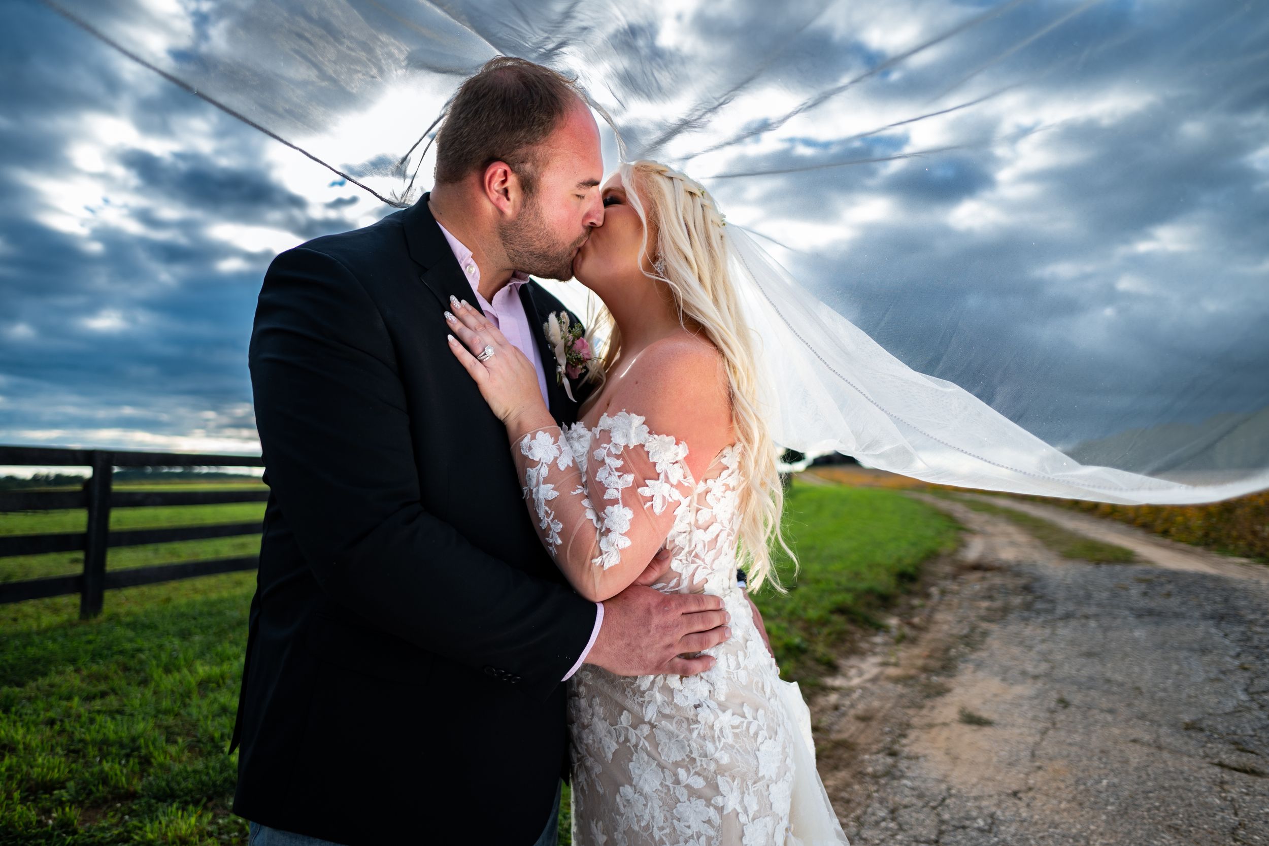 Dramatic wedding portrait with sweeping veil against stormy sky and rustic fence.
