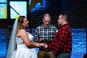 Wedding ceremony with couple in plaid shirts and minister in rustic church setting.