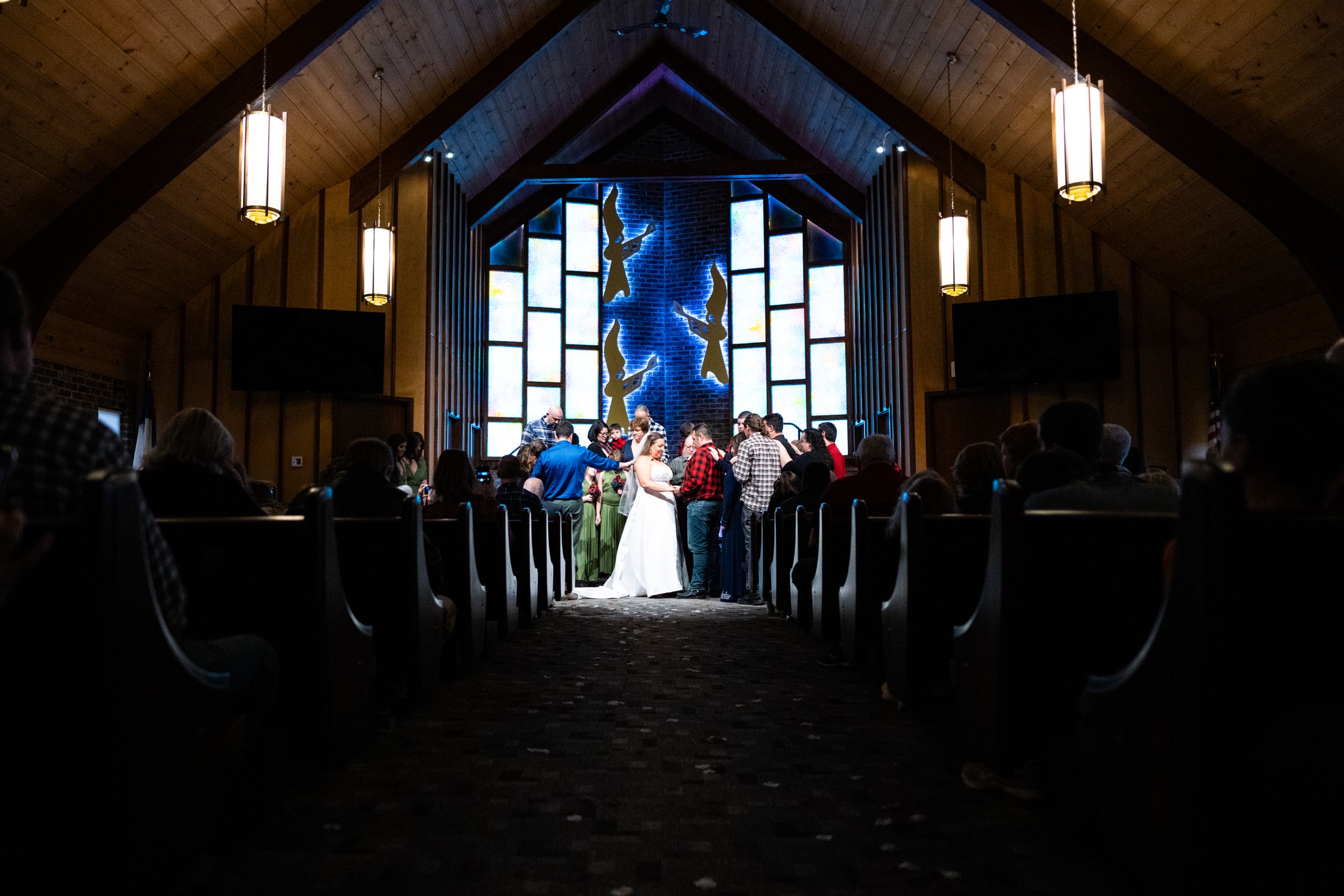Dramatic wide shot of wedding ceremony in chapel with illuminated stained glass windows.