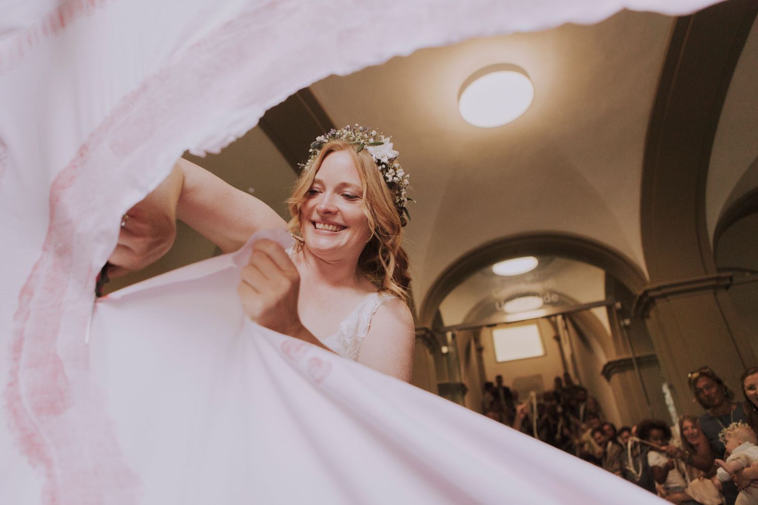 Behind flowing fabric, a joyful bride in a crown stands beneath arched hallway ceilings at her wedding.