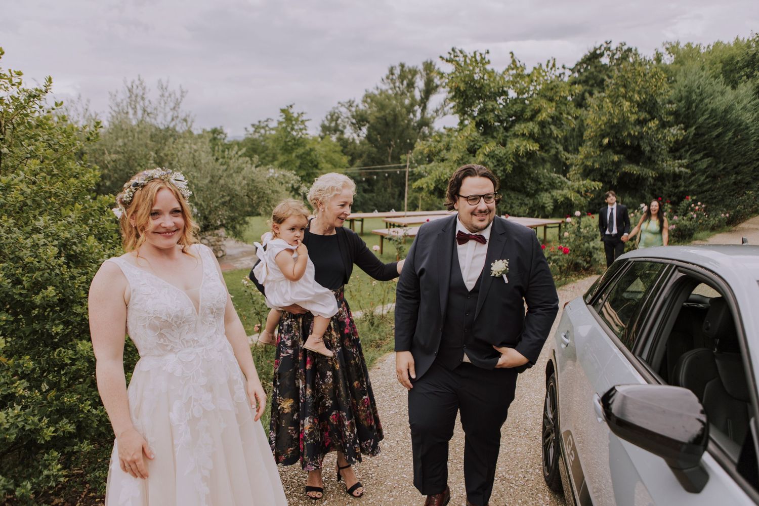 Wedding party gathered around a parked car on a tree-lined path.