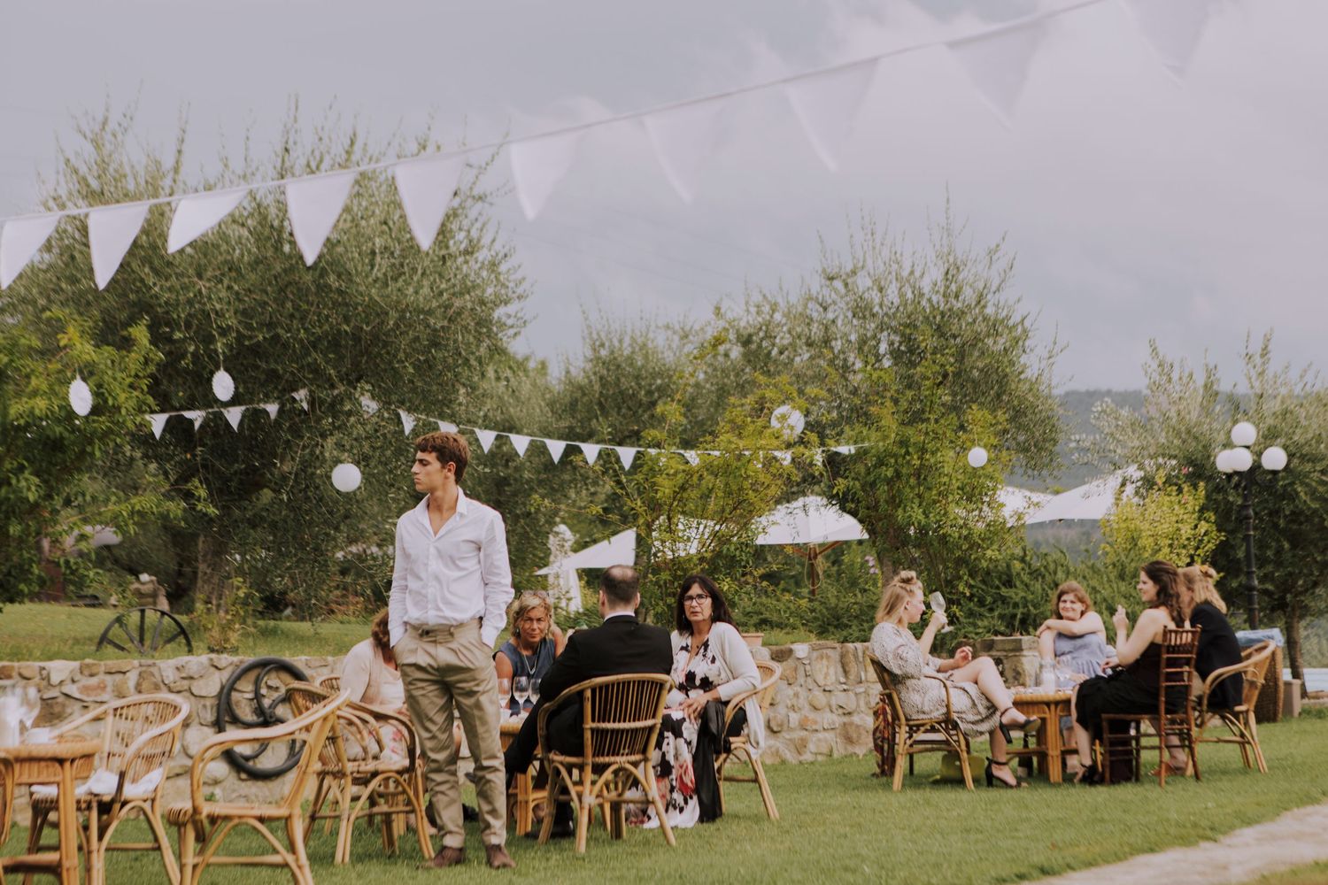 An outdoor wedding reception with white bunting decorations and guests seated at rustic tables.