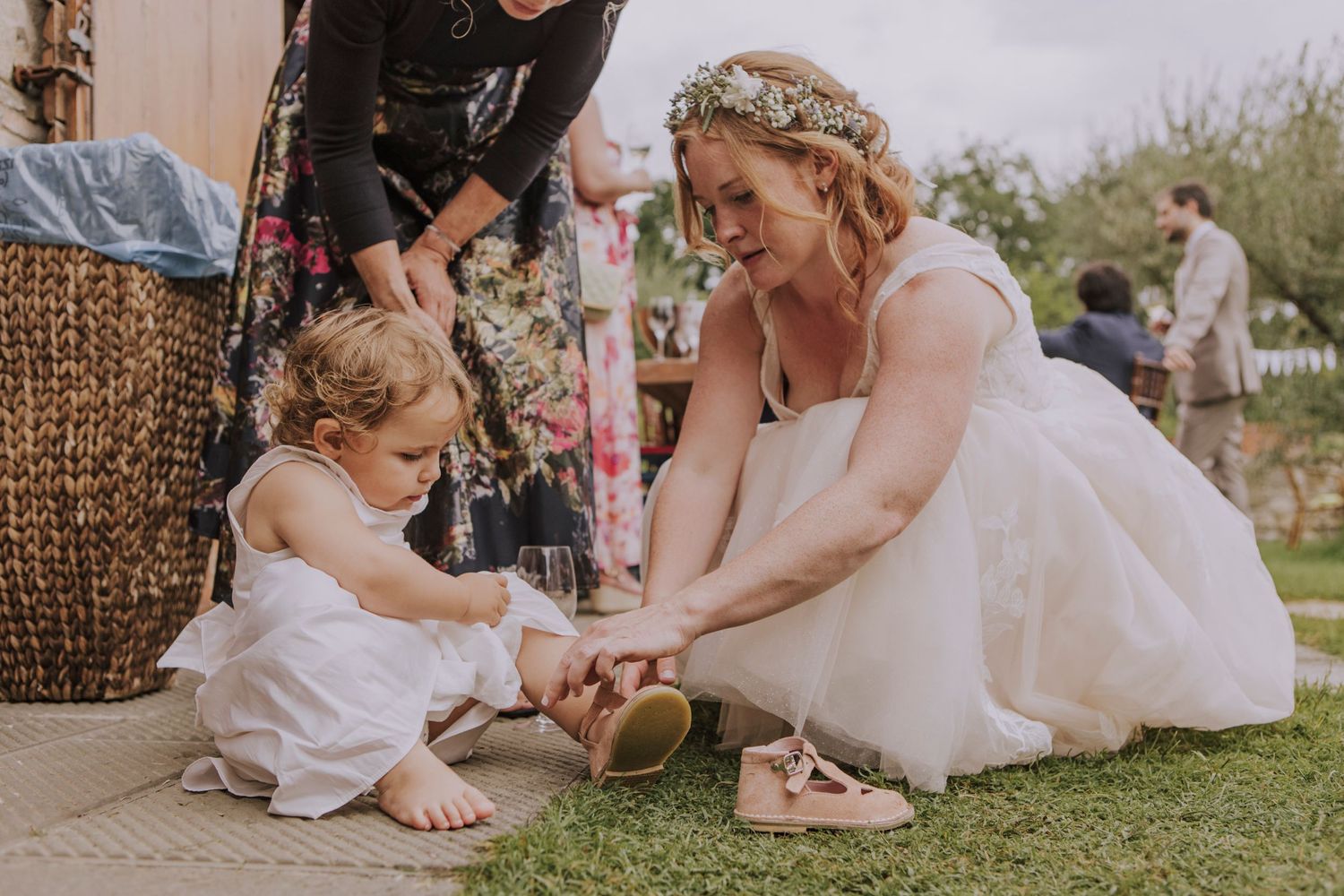 A tender moment shared between a woman in a white wedding dress and small child sitting on grass outdoors.