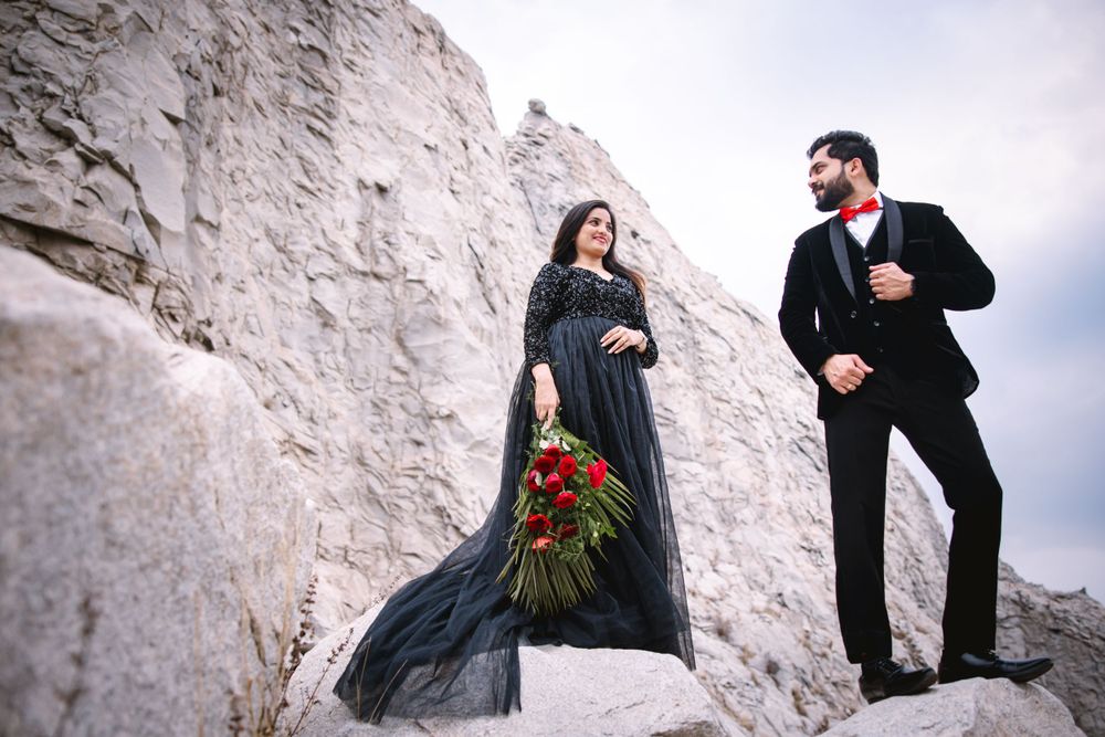 Couple in black formal attire poses against white stone wall for romantic wedding photos.
