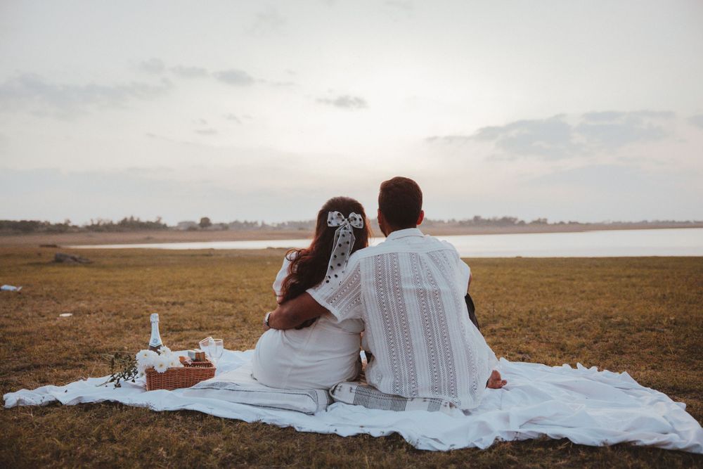A couple sitting on a white blanket by the waterfront during golden hour.