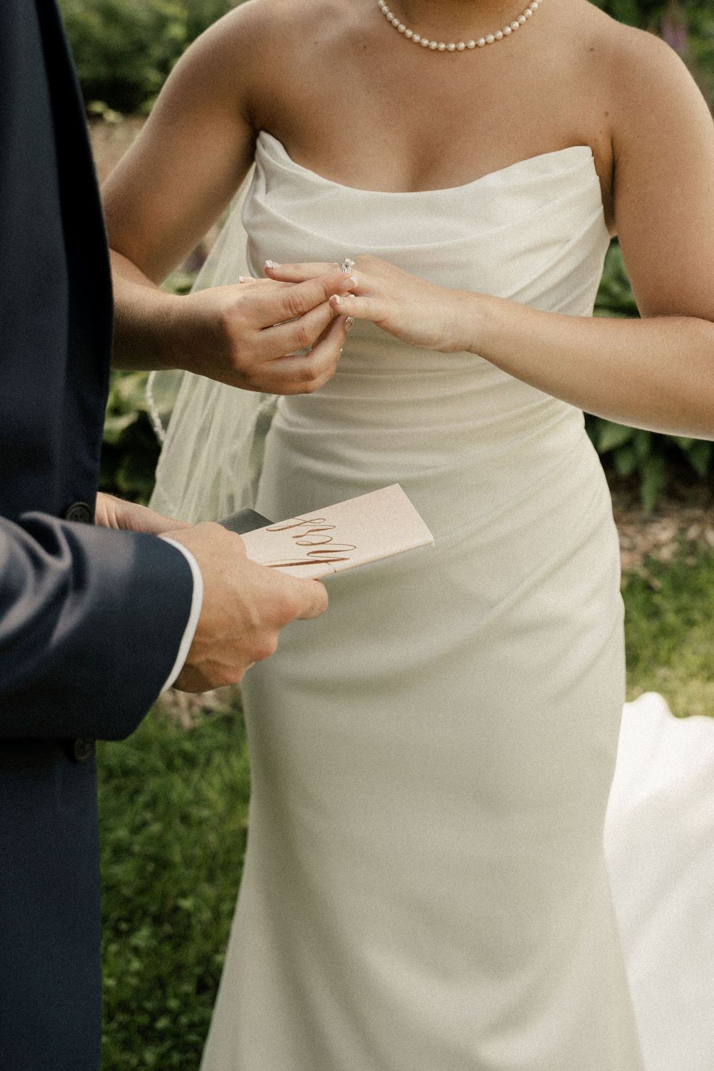 Close-up of wedding ceremony showing hands exchanging vows with white dress and pearl necklace details.