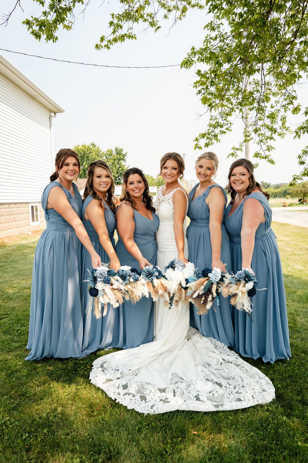Bridesmaids in dusty blue dresses gather around a bride's lace wedding gown train on green grass.