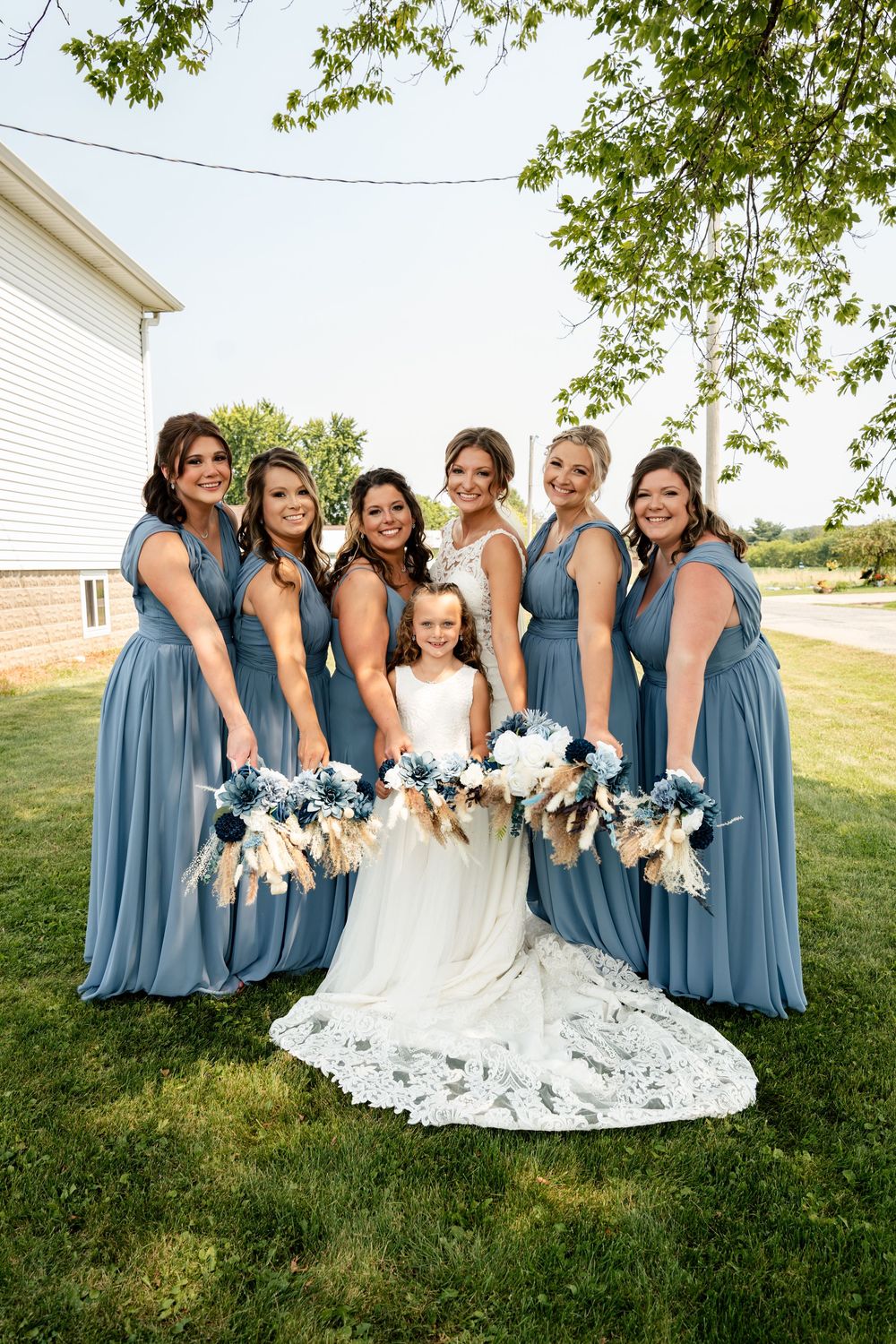 Wedding party in coordinated dusty blue dresses poses together on a sunny lawn.