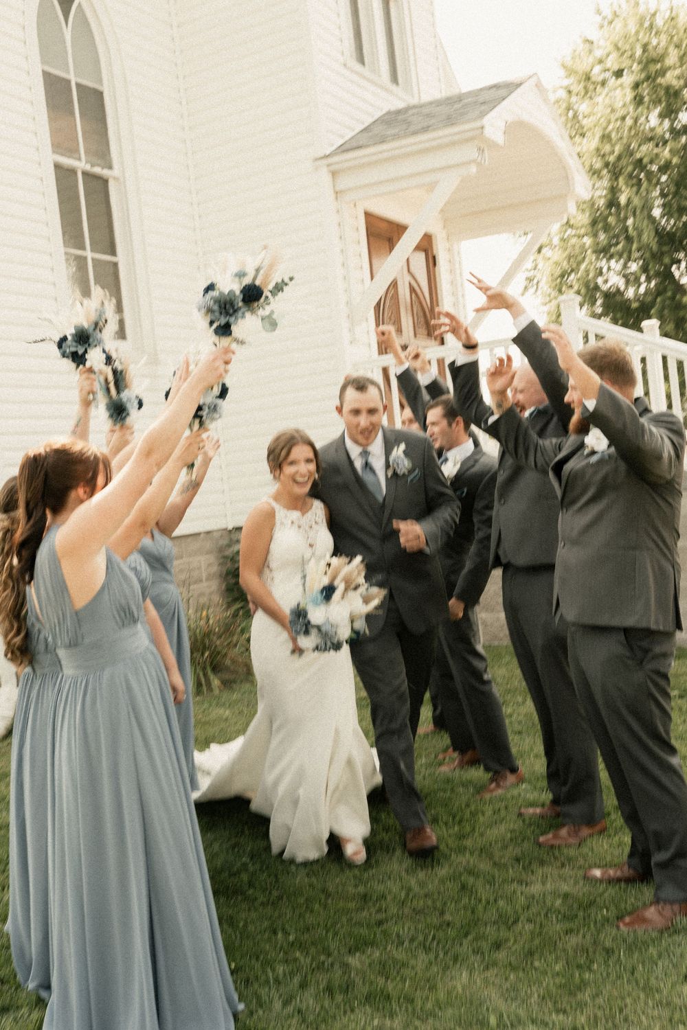 Wedding party in blue dresses and dark suits poses energetically on church lawn.