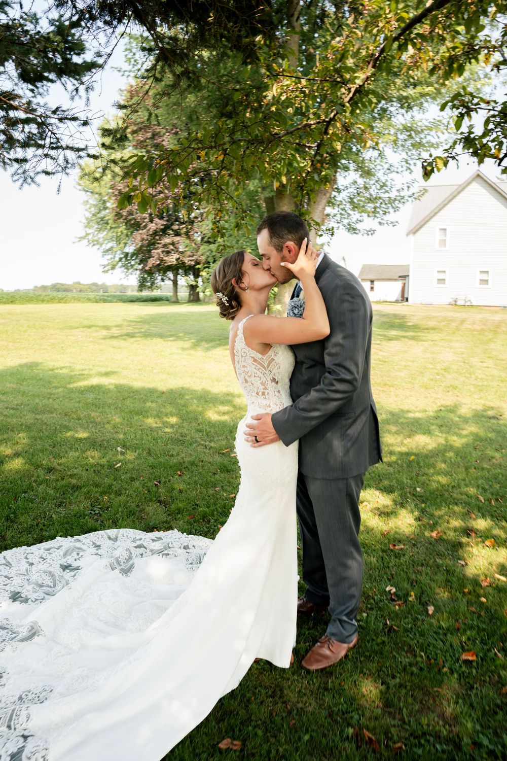 A romantic wedding photo of a couple embracing under a tree on a sunny day, with the bride's dress trailing on the grass.