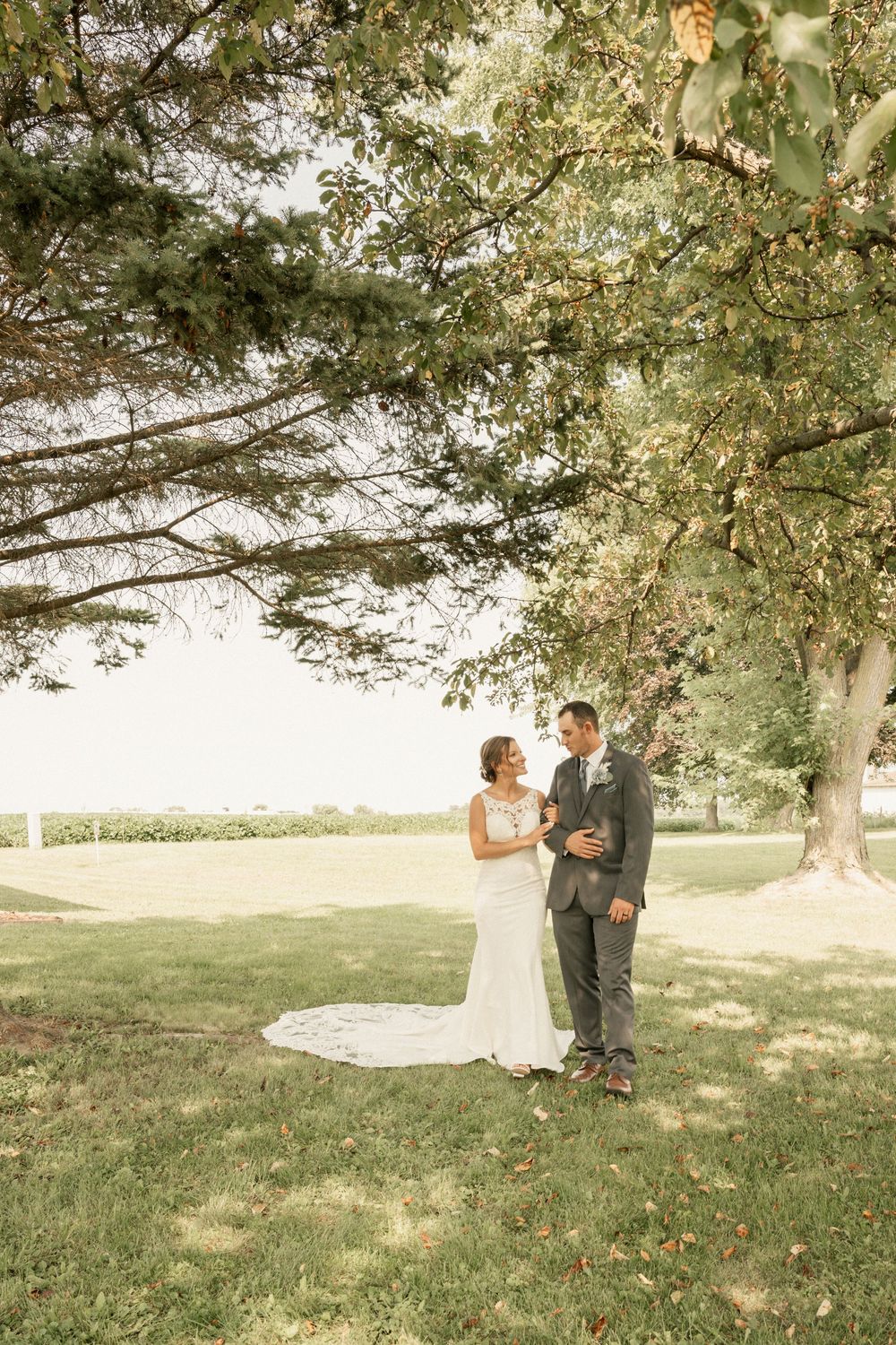 A sunlit outdoor wedding portrait showing a couple standing together on a grassy hill with trees in the background.