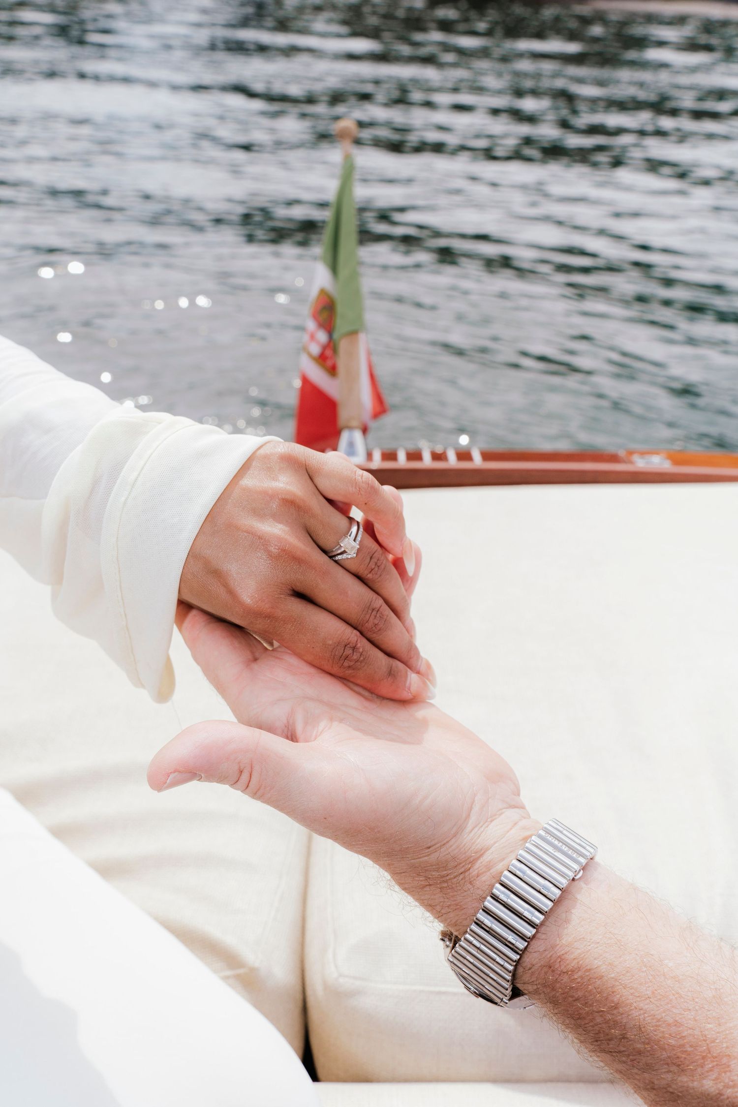 A hand wearing an engagement ring is shown against black fabric with pink flowers in soft focus.