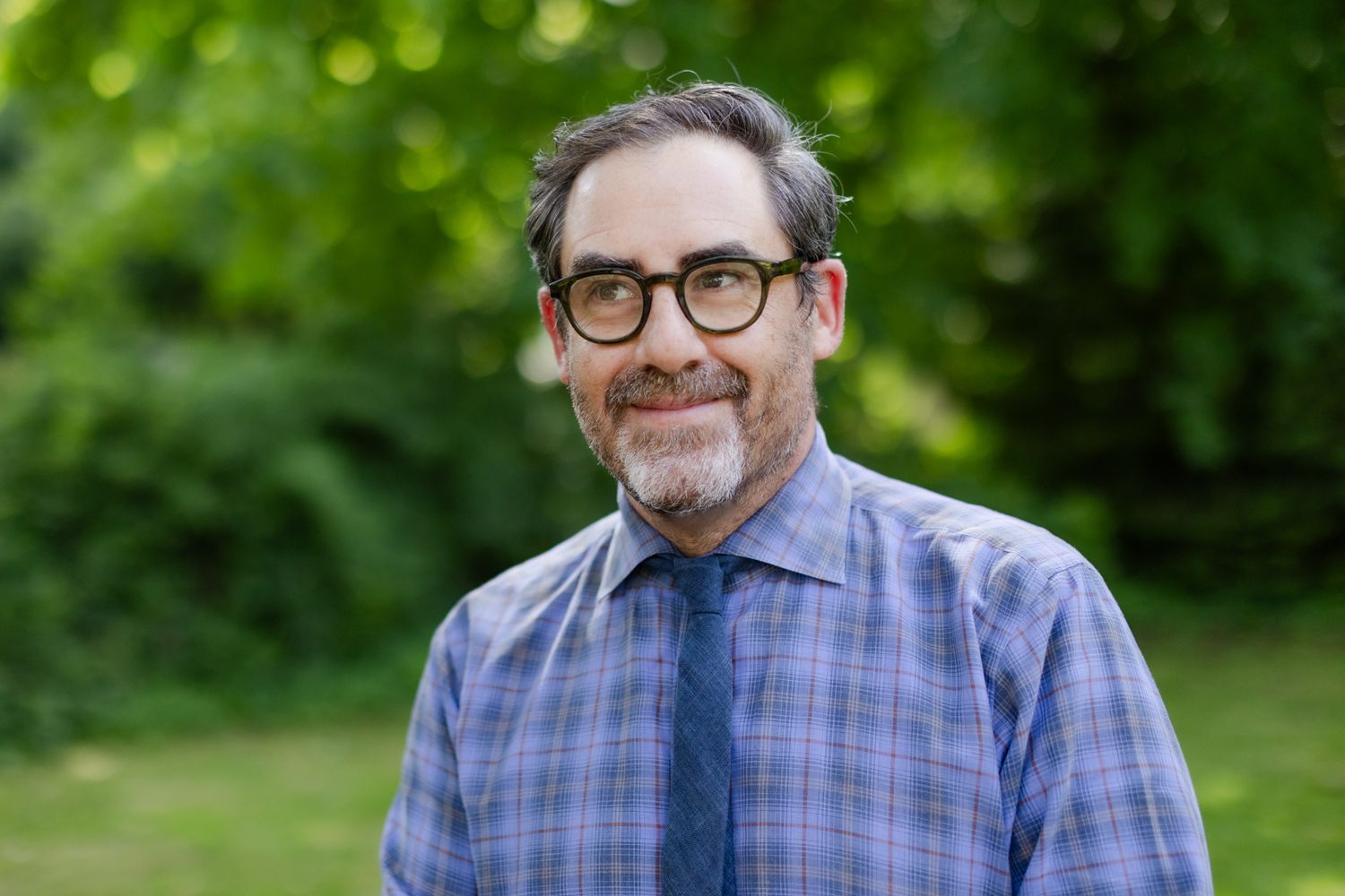 Professional wearing glasses and a plaid button-up shirt smiling outdoors with green foliage background.
