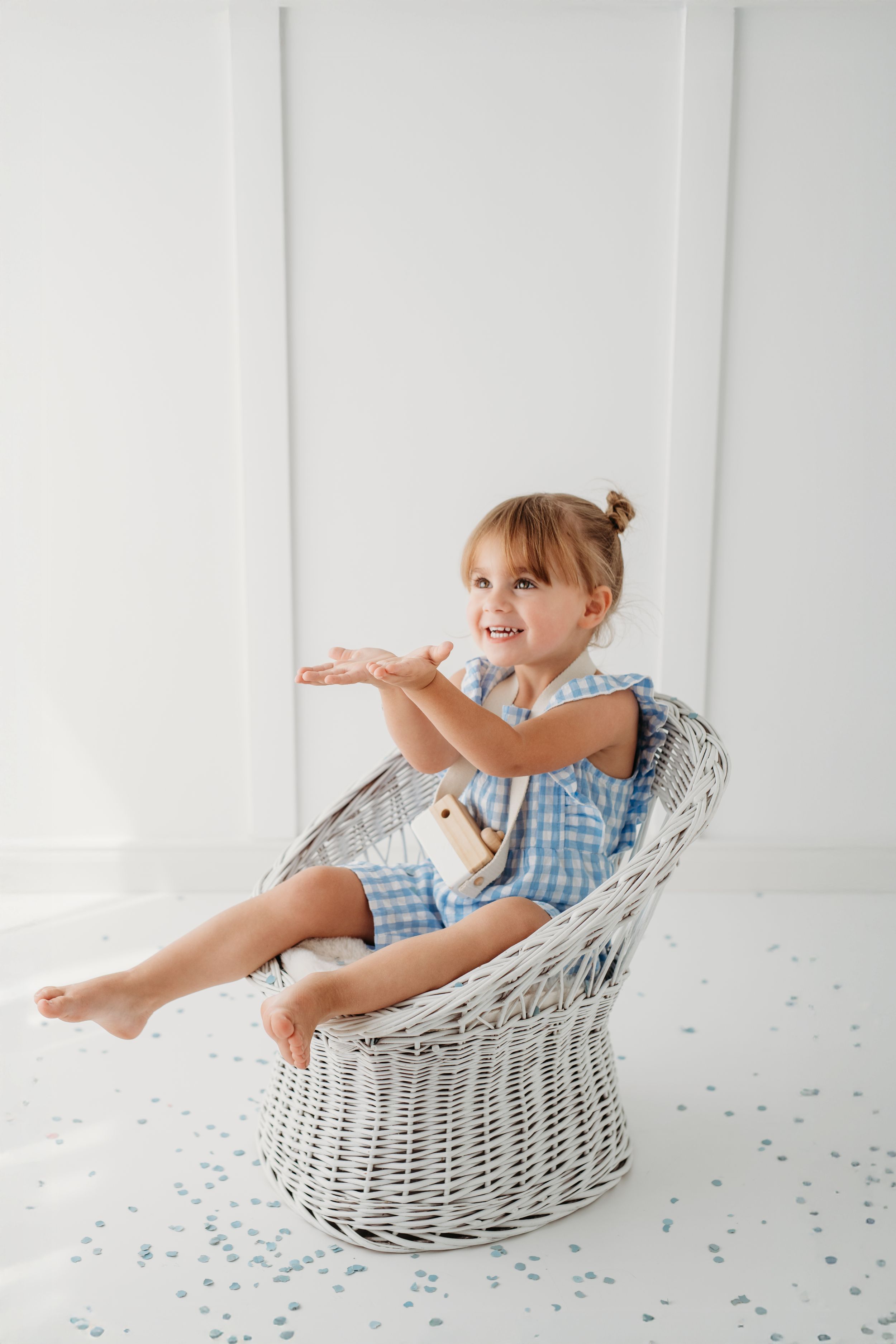 A cheerful young girl in a blue dress sits in a white wicker chair, smiling and gesturing with her hands.
