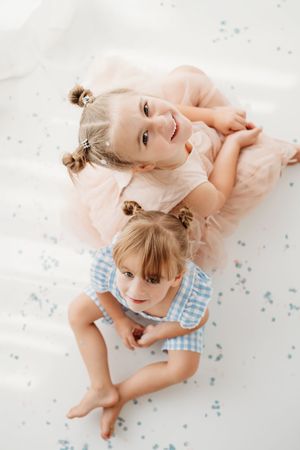Two young sisters in pink and blue outfits lying on a white floor, smiling up at the camera.