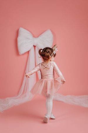 Young dancer in pink leotard and tutu performs ballet pose against coral wall with large white bow decoration.