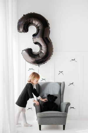 Child plays with teddy bear in grey armchair under large silver number 3 balloon against white wall with plane decals.