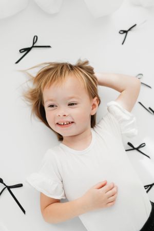 A young child in a white t-shirt lies on a white surface decorated with black bow patterns, smiling brightly.