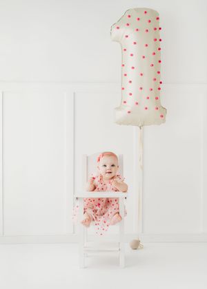 Baby girl in pink polka dot dress sits in white chair beside a number 1 balloon for her first birthday.