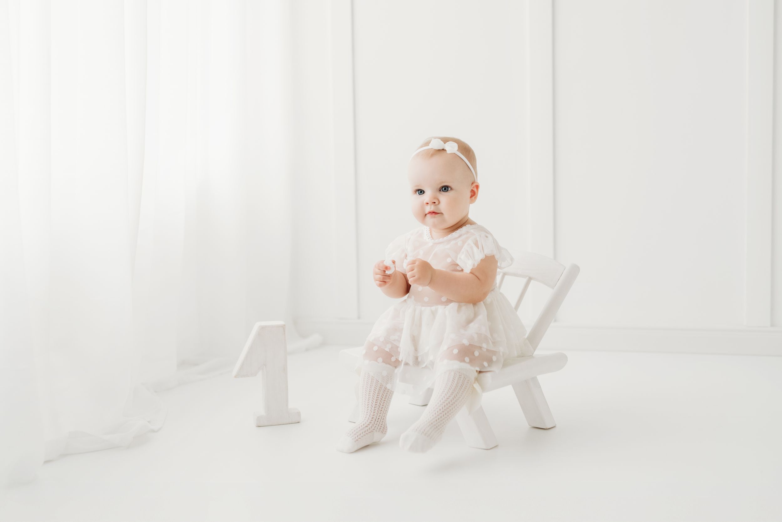 Baby girl in white lace dress sits on small white chair with number one decoration for first birthday photo.