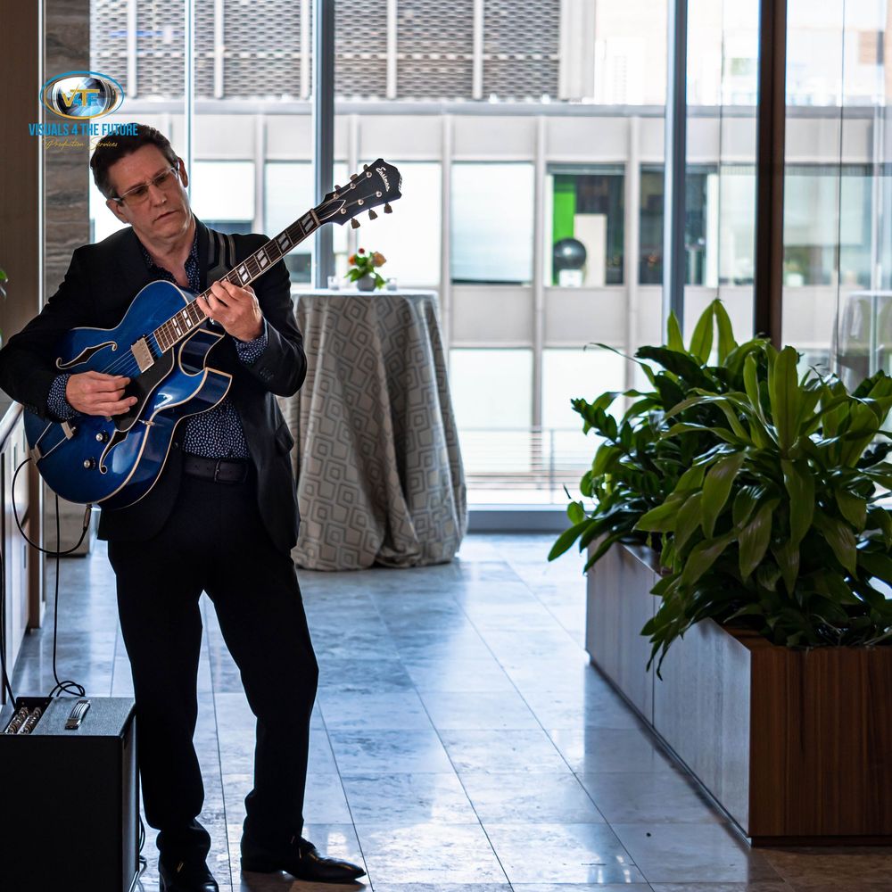 Musician plays a blue electric guitar while seated in a modern office hallway with large windows and indoor plants.