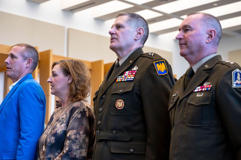 Military service members and civilians stand in a row watching an event with focused attention.