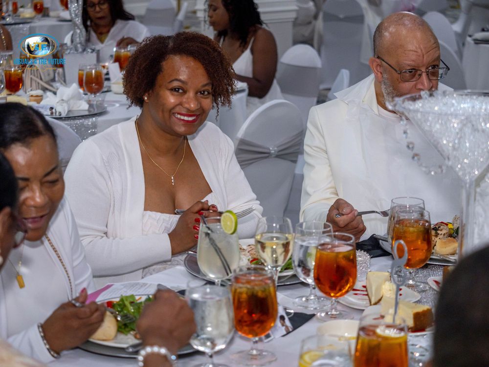Guests in white attire gather around tables at a formal dining event with iced tea drinks and place settings.
