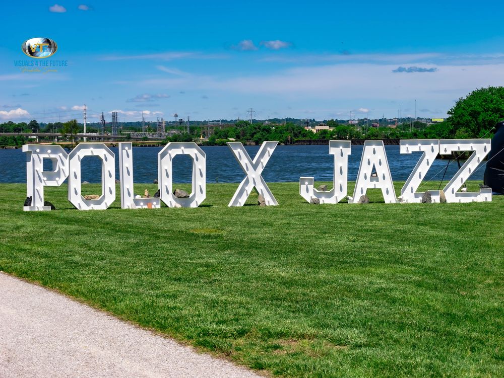 Large white letters spelling 'POLO X JAZZ' displayed on green grass with trees and buildings in the background.