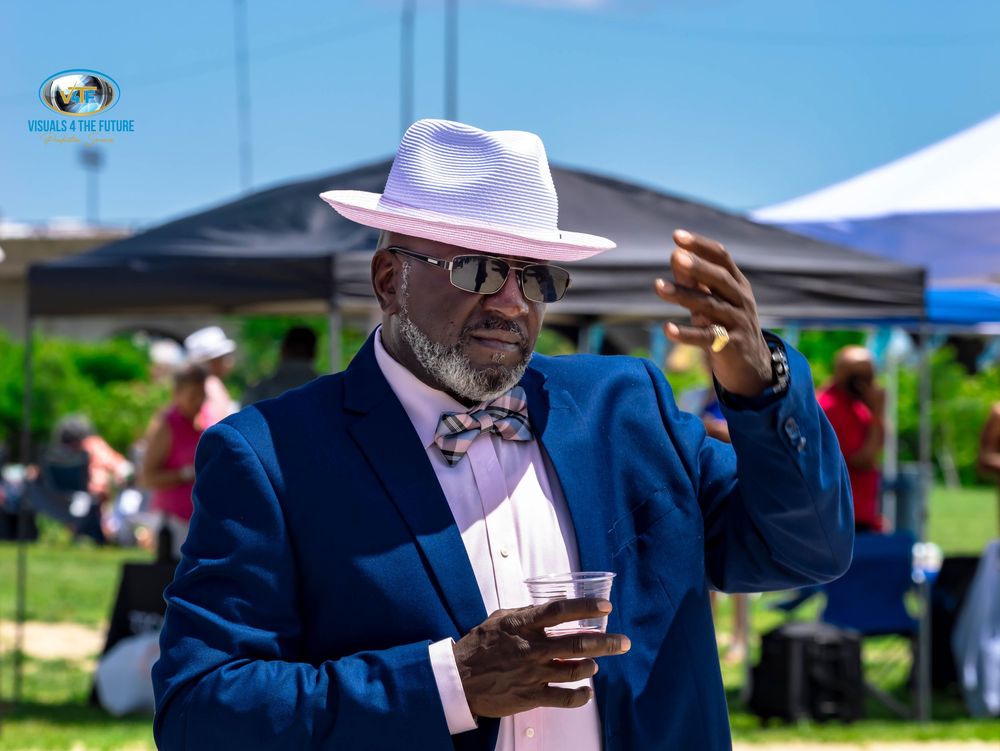 People in formal attire enjoy drinks and socialize at an outdoor event under blue tents and umbrellas.
