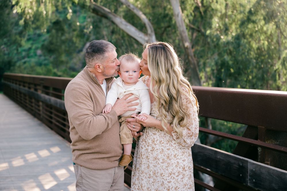 A family shares tender moments together on a wooden bridge surrounded by lush greenery on a sunny afternoon.