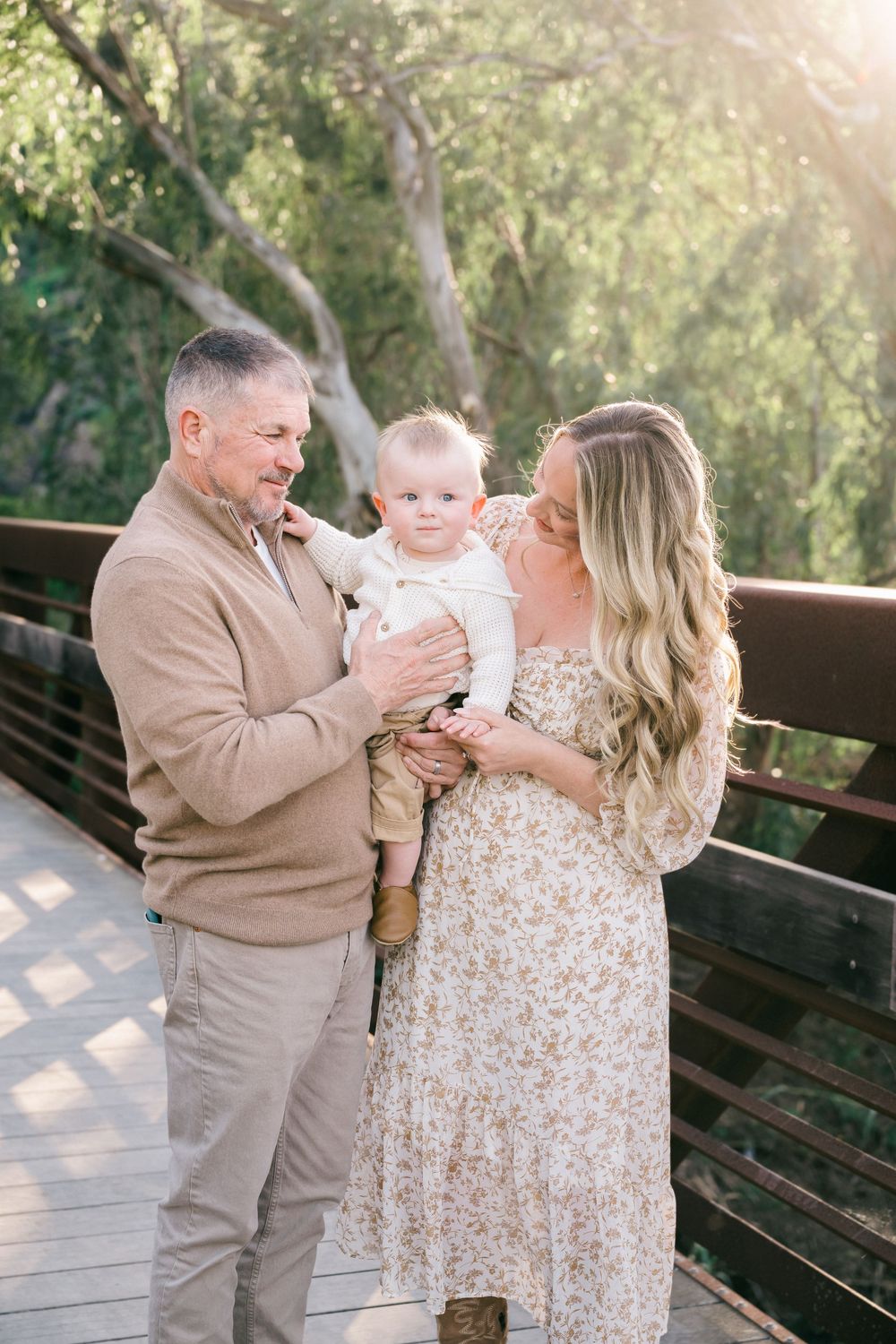 A family shares tender moments together on a wooden bench in a sunlit outdoor setting with trees in the background.