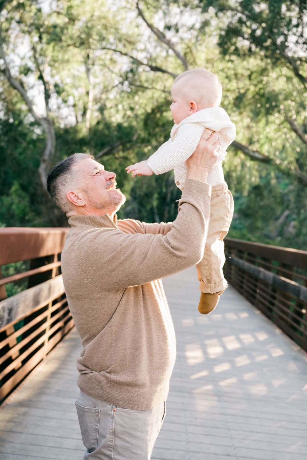 Four sequential photos showing someone in a beige sweater joyfully lifting and playing with a small baby on a wooden deck.