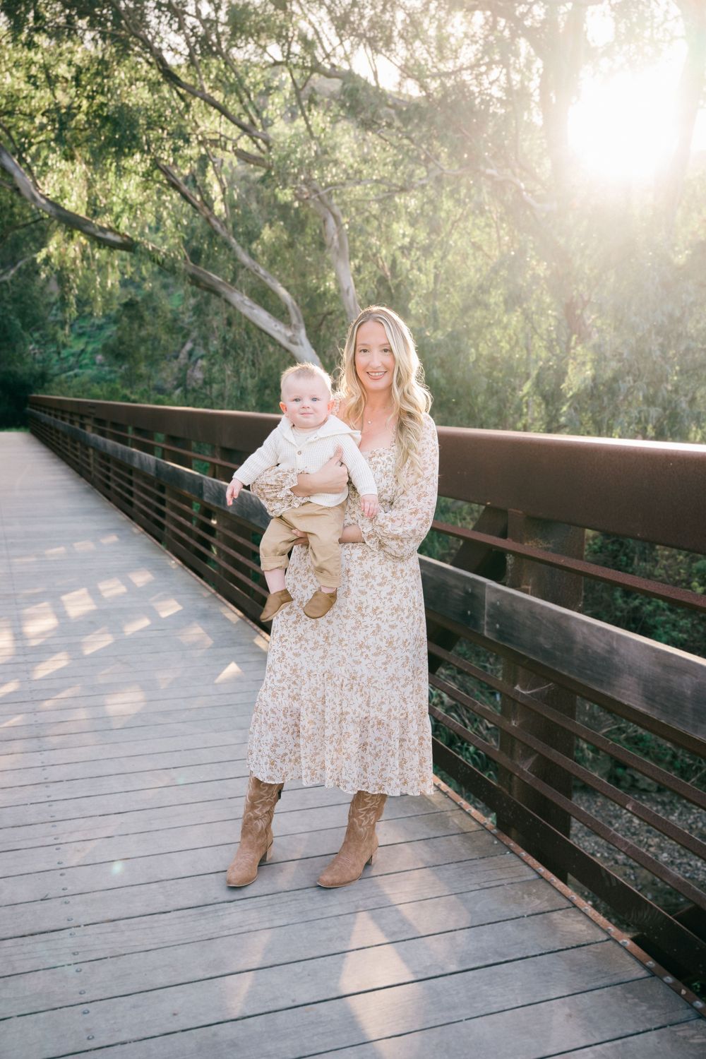 A mother and baby share tender moments on a wooden boardwalk surrounded by sunlit trees during golden hour.