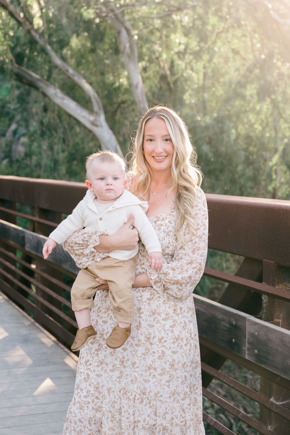 A mother and baby pose together on a wooden bridge in a sunlit forest setting wearing coordinating light colored outfits.