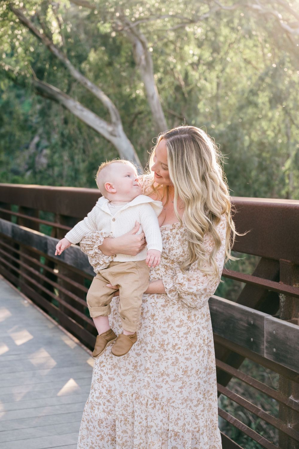 Two people in coordinating cream and floral outfits share a tender moment on a wooden bridge in a natural setting.