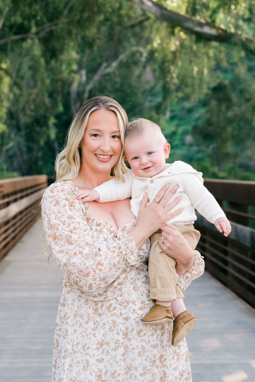 A mother and baby share joyful moments together on a wooden bridge surrounded by lush green trees.