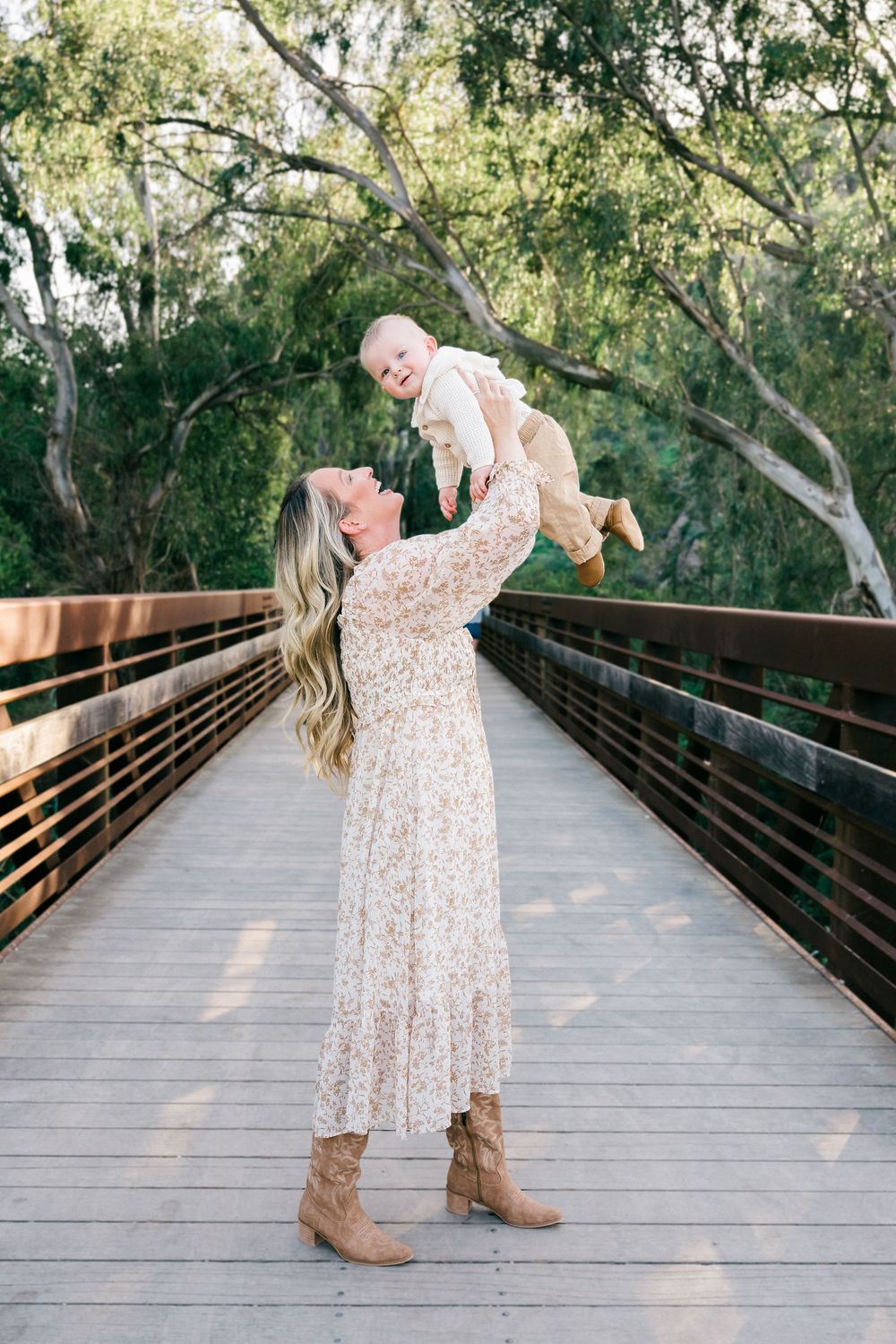 A joyful moment captured on a wooden bridge as a woman in a floral dress lifts a baby against a natural backdrop.