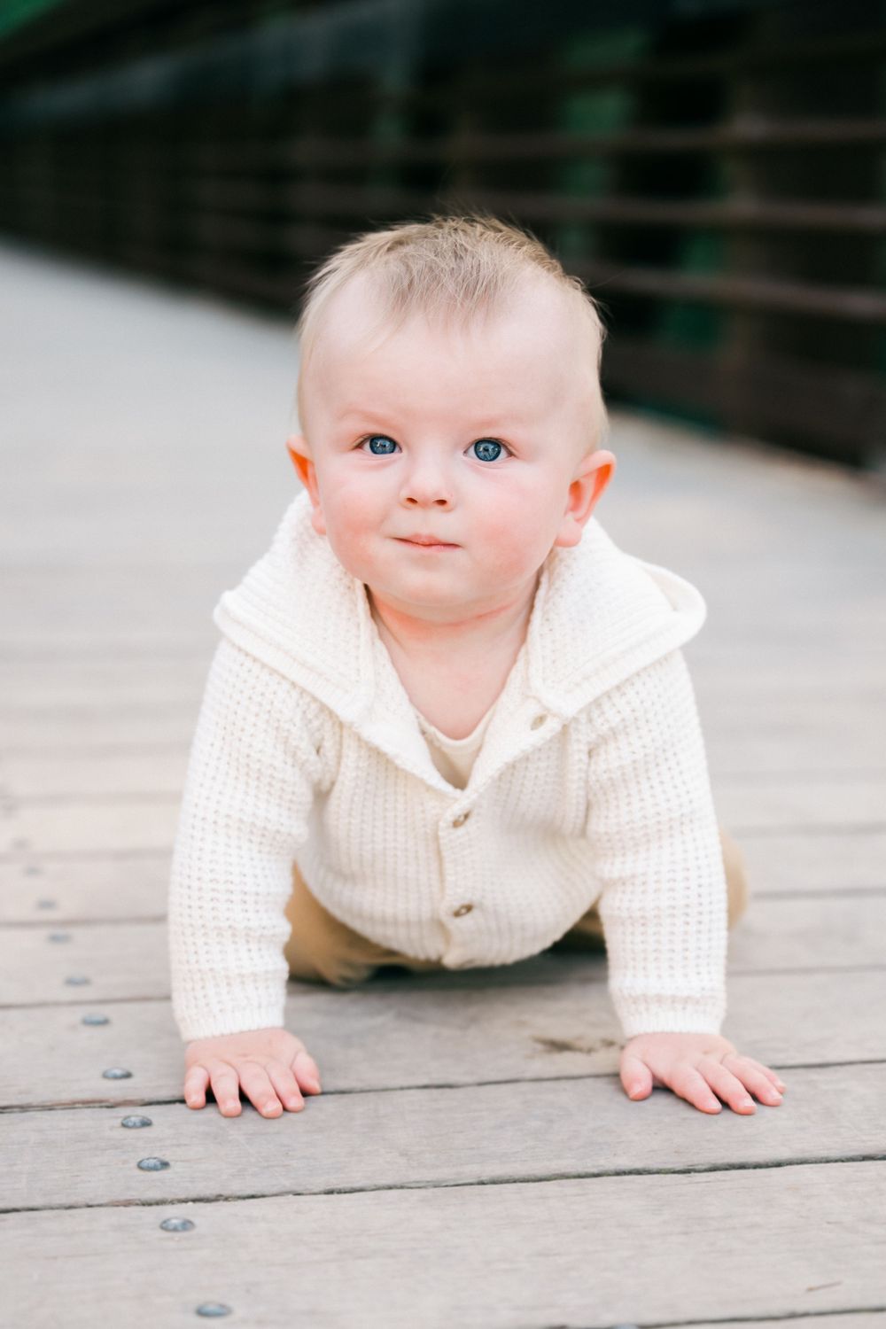 A baby in a white sweater crawls on wooden decking while looking at the camera.
