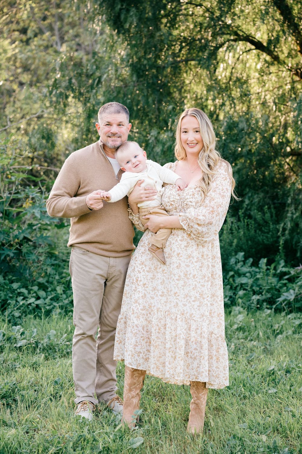 A family poses together outdoors on a grassy hill with willow trees in the background on a sunny day.