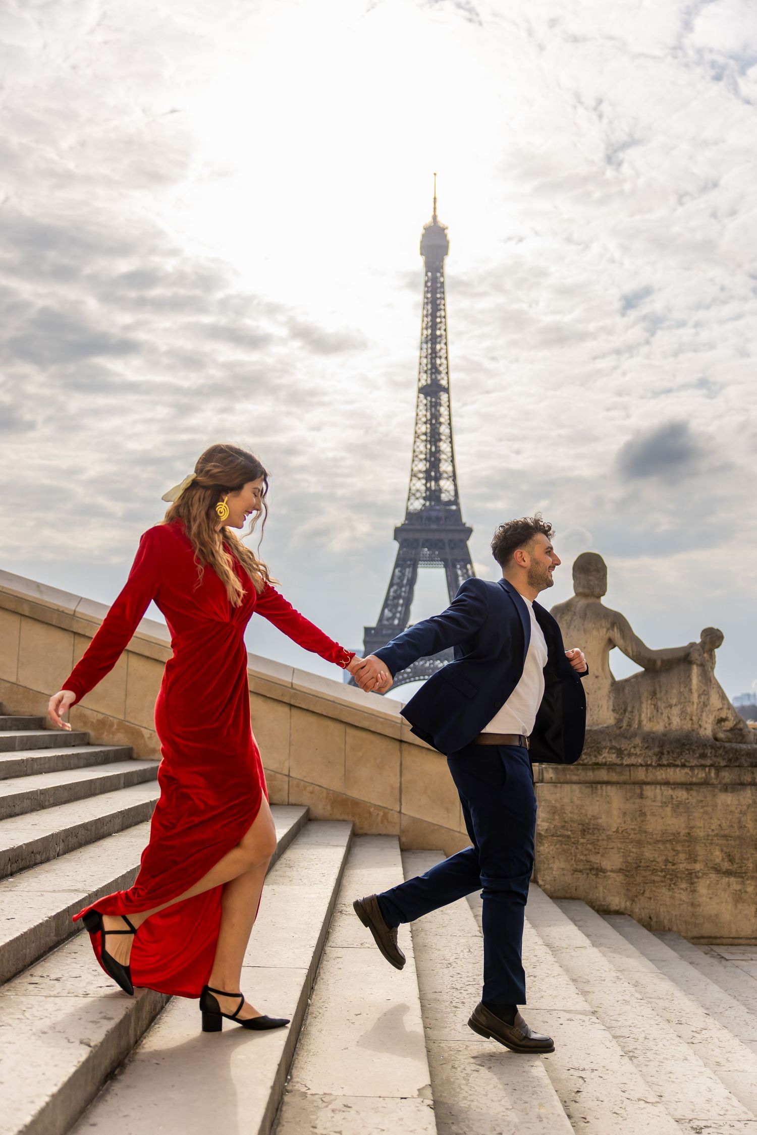 A couple in formal attire playfully descends steps near the Eiffel Tower in Paris on a cloudy day.