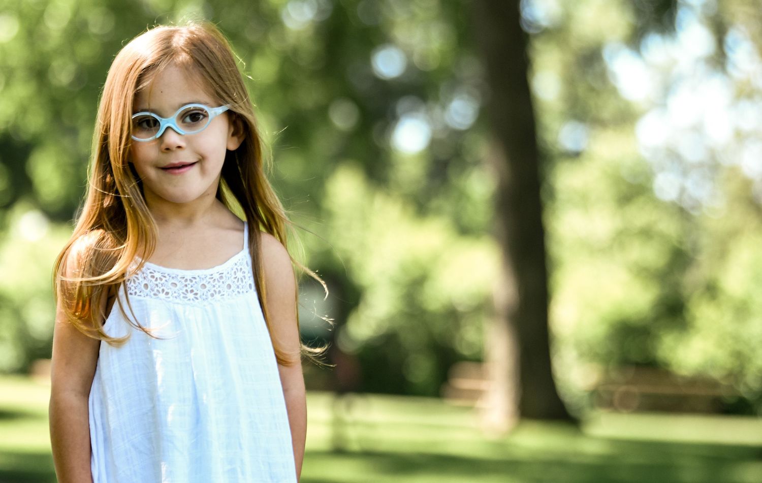 Young child wearing a white summer dress stands outdoors in a sunny park setting.