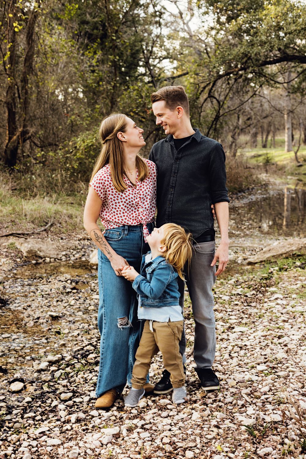 A young couple shares a tender moment with their child while standing on fallen autumn leaves in a wooded area.