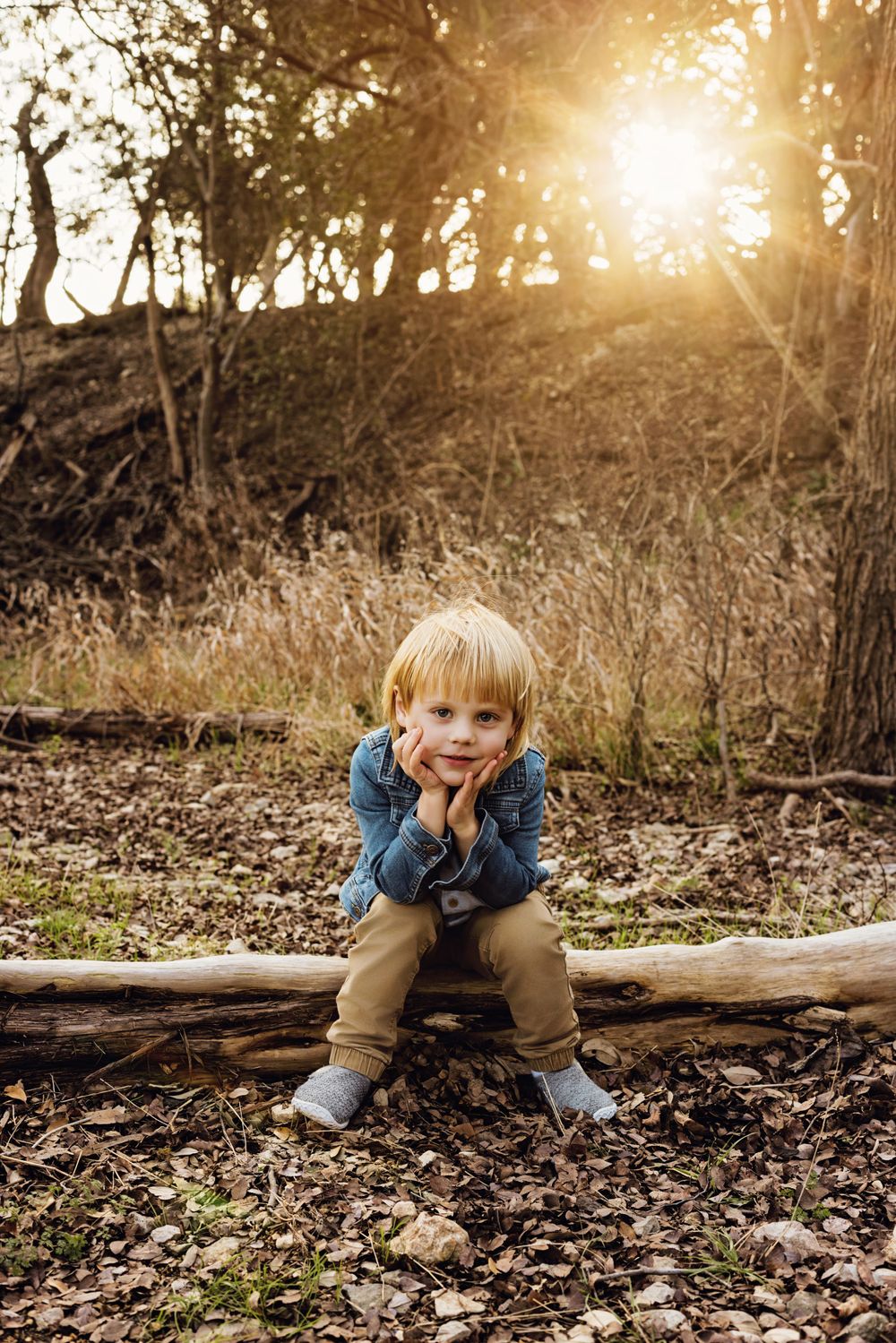 Young child sits on fallen log in woods during golden sunset wearing blue sweater and tan pants.