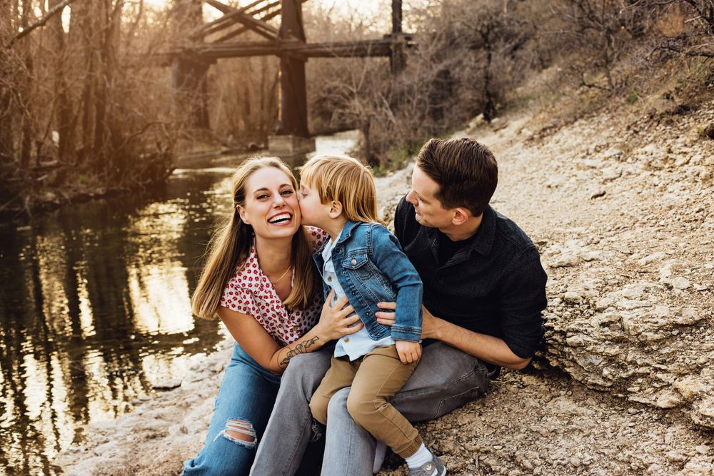 A family sits together by a quiet stream at sunset with a rustic bridge in the background.