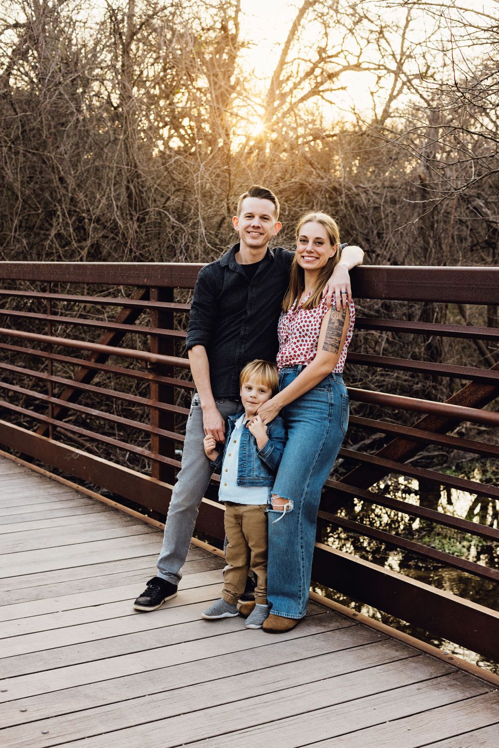 A family stands together on a wooden bridge during sunset with warm golden light filtering through trees in background.