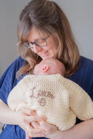 New mum holding newborn baby on her front. Newborn baby is wearing a handmade knitted cardigan with name on.