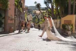 A colourful shot of a newlywed couple walking across a street in Italy, with crowds around them. The brides veil is dragging behind her.